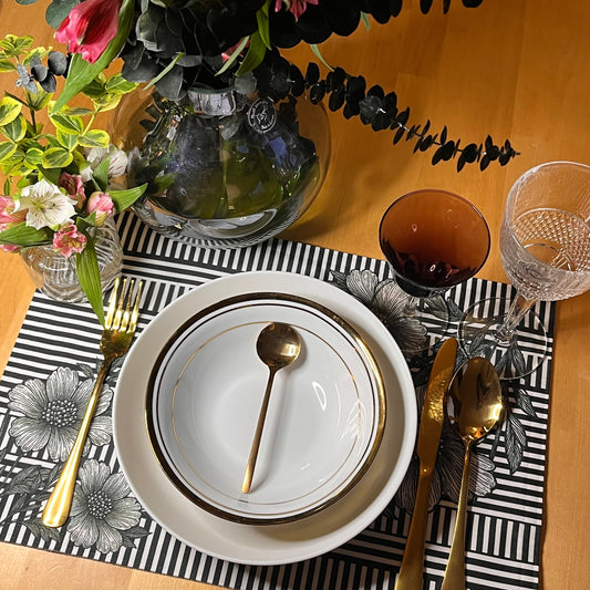 Black and white floral placemat with engraved-style flowers over graphic stripes on a dining table setting with white plates, gold cutlery, and floral decorations.