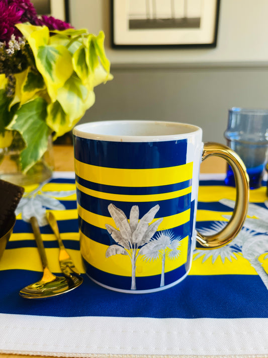 A ceramic mug with a navy blue and yellow striped pattern, featuring white palm tree illustrations and a shiny gold handle, set on a matching blue and yellow placemat.