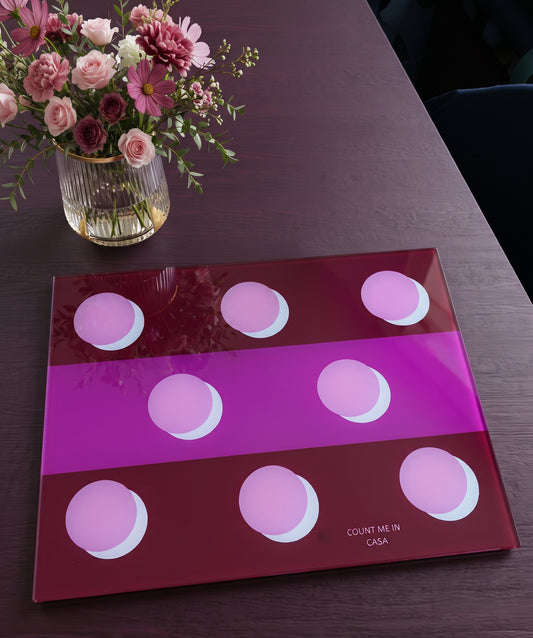 A glossy rectangular glass tray with a geometric design of pink and purple circles, placed on a dark wooden table next to a glass vase of flowers.