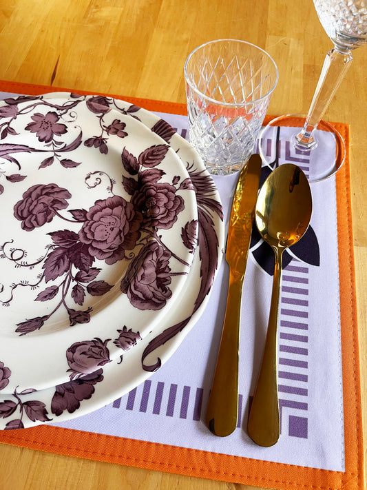 Table setting with floral plate, gold cutlery, and clear glass on an orange placemat, zoom
