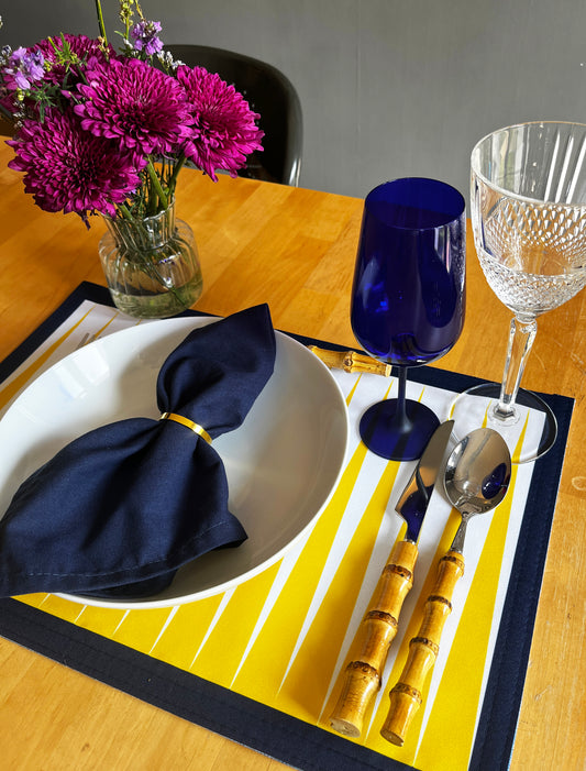 A styled table setting featuring a yellow and white striped placemat with a navy blue border, a white bowl with a navy blue napkin, gold-handled cutlery, a cobalt blue tumbler, a clear wine glass, and a vase of magenta flowers on a wooden table.