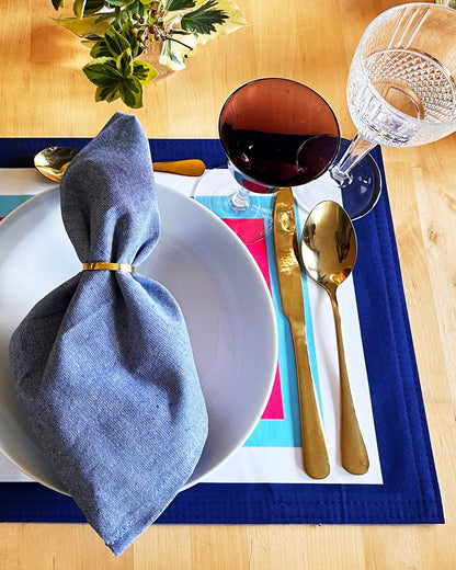 Dining table setting with a blue napkin, white plate, gold cutlery, and colorful placemat.
