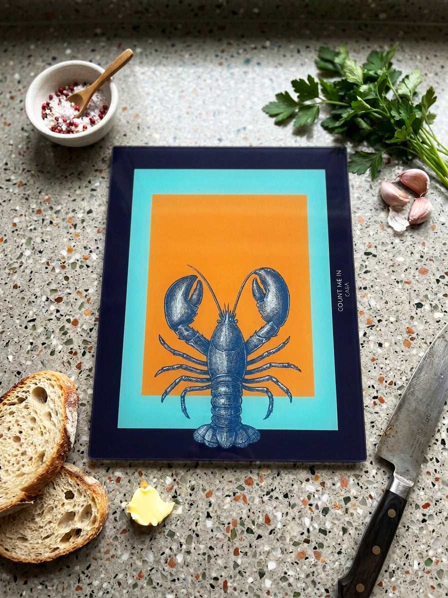 A rectangular art print featuring a blue lobster illustration on an orange background with turquoise borders, placed on a speckled countertop with bread, garlic, parsley, a bowl, and a knife around it.