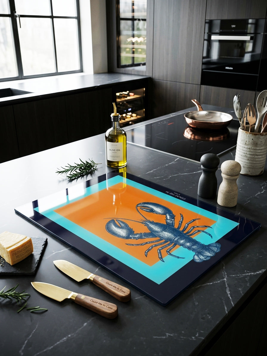 A rectangular cutting board with a dark navy border and a bright orange and turquoise center featuring a blue lobster illustration, styled on a dark kitchen island with cheese, knives, olive oil, and kitchen accessories.