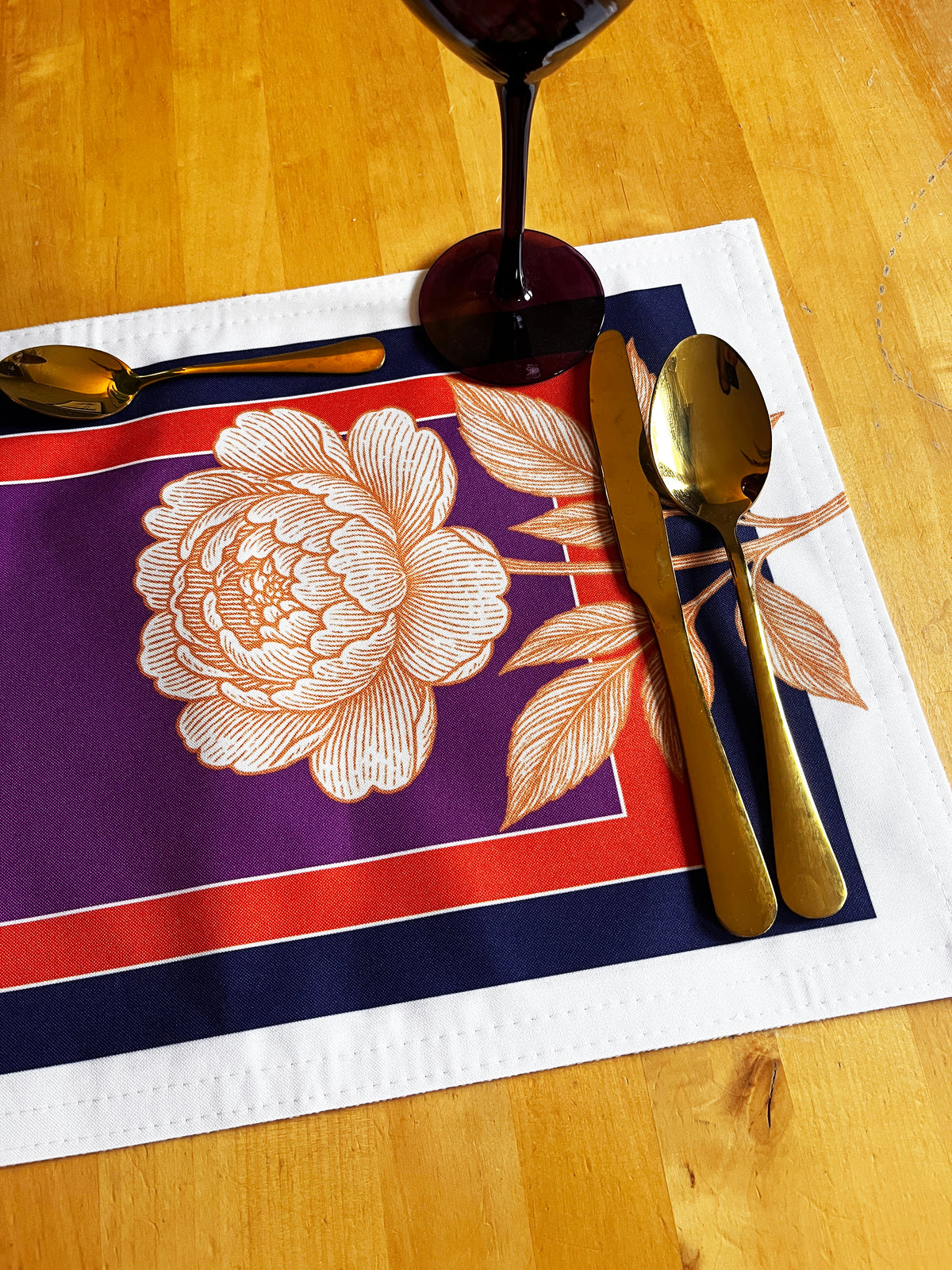 A rectangular table placemat with a purple center, red border, navy outer border, and a large cream floral print, styled on a wooden table with gold-toned cutlery and a wine glass base visible.