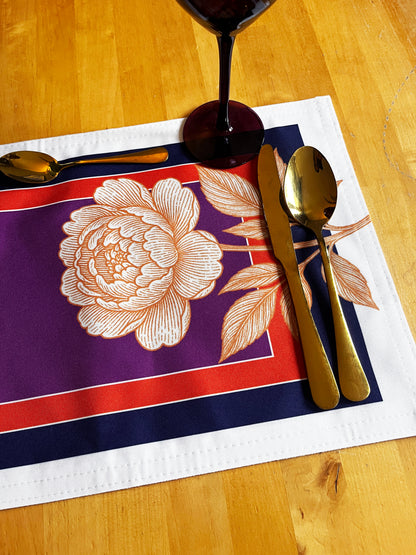 A rectangular table placemat with a purple center, red border, navy outer border, and a large cream floral print, styled on a wooden table with gold-toned cutlery and a wine glass base visible.