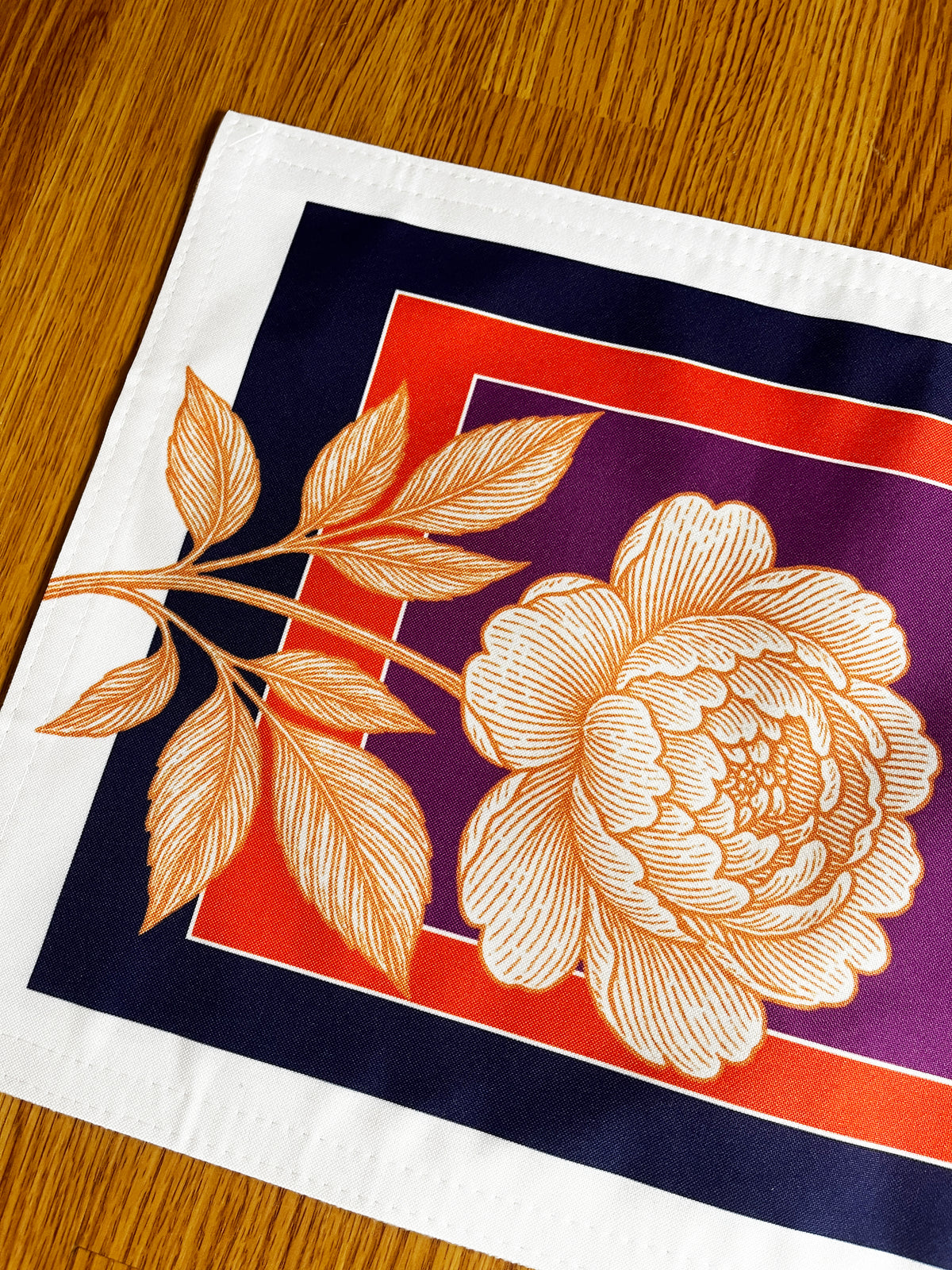 A square white fabric napkin or placemat featuring a large cream-colored peony flower and leafy branch over a geometric background of purple, red, and navy blue, laid flat on a wooden table.