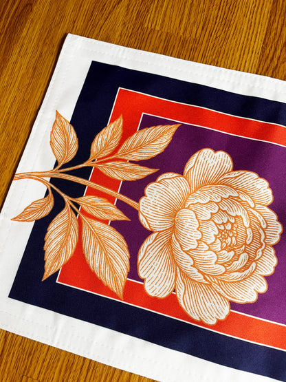 A square white fabric napkin or placemat featuring a large cream-colored peony flower and leafy branch over a geometric background of purple, red, and navy blue, laid flat on a wooden table.