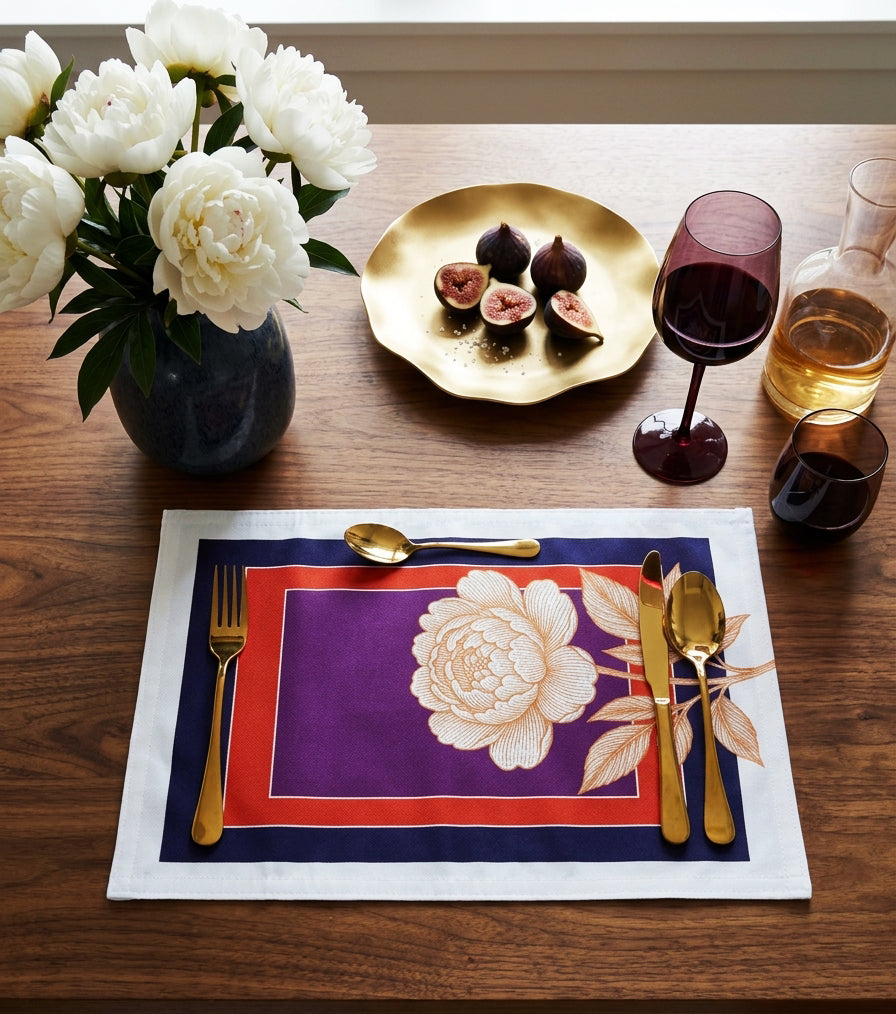 A styled dining table setting featuring a rectangular linen placemat with a purple center, a large white peony illustration, and navy and white borders, arranged with gold-toned flatware and surrounded by a vase of white flowers, a gold plate with figs, and wine glasses.