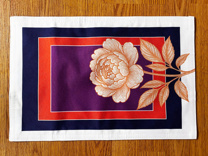 A square fabric placemat with a white border, featuring a central design of a peach-colored peony flower and leaves over concentric purple, red, and navy blue rectangles, laid flat on a wooden table.