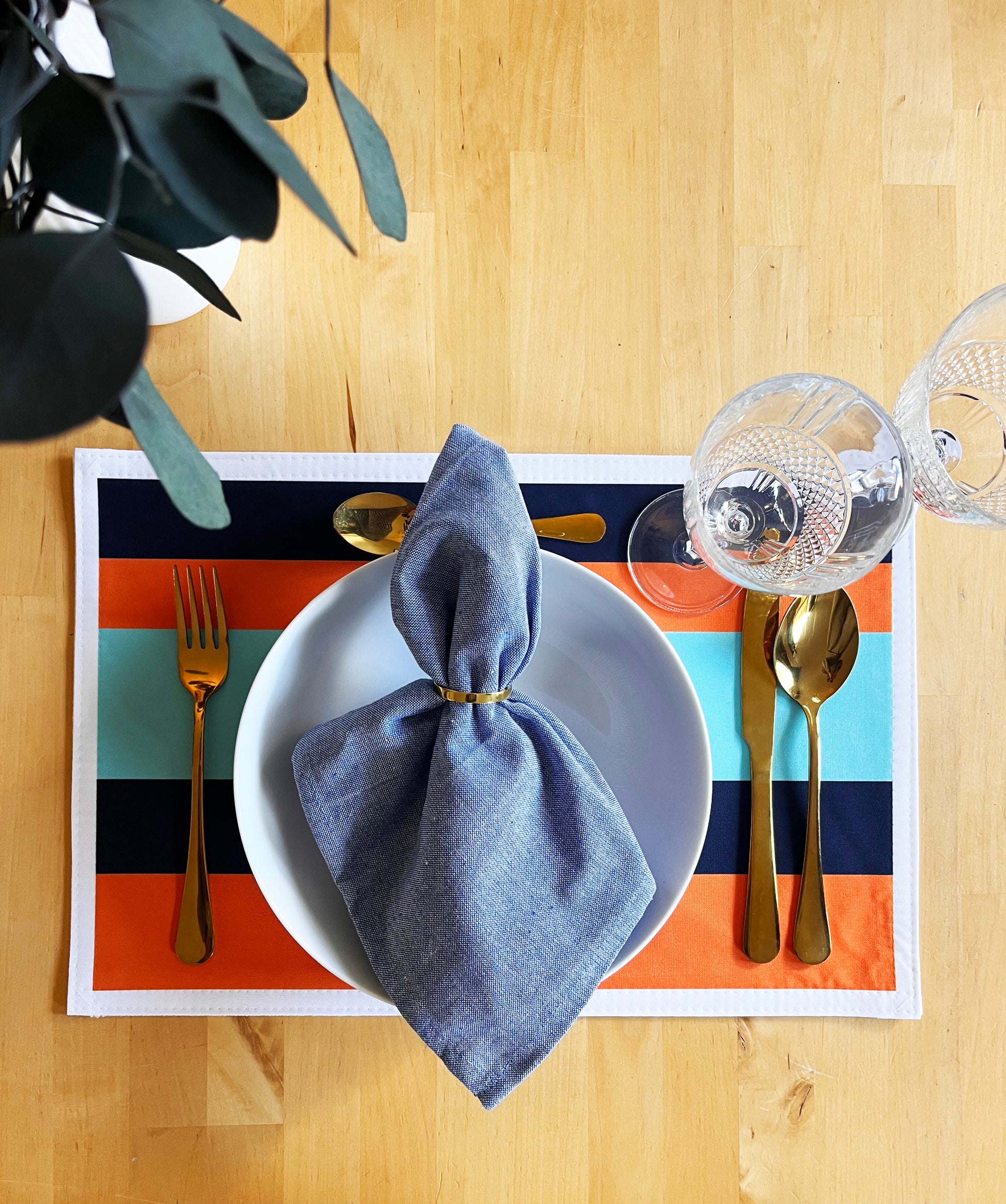 Placemat with striped pattern, designed in the UK, white plate, blue napkin, gold cutlery on wooden surface.