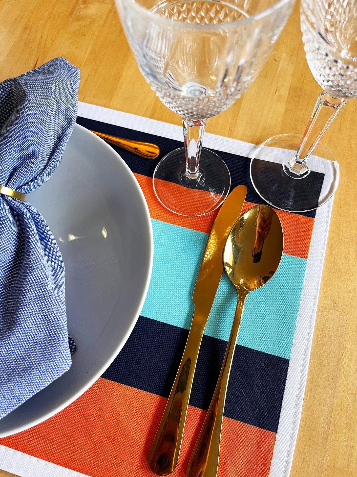 Dining table setting with gold cutlery, blue napkin, and clear glasses on a colorful exclusive design placemat.