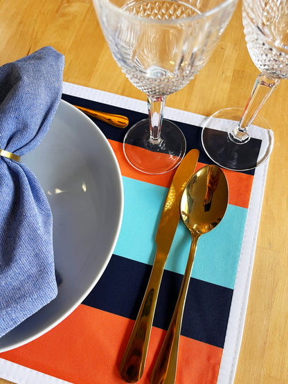 Dining table setting with gold cutlery, blue napkin, and clear glasses on a colorful exclusive design placemat.