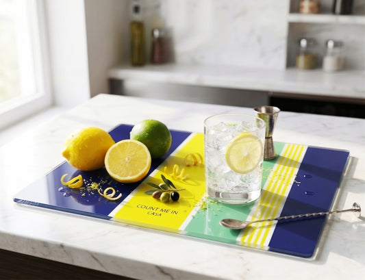 A square glass serving board with a colorful geometric pattern of navy blue, yellow, and mint green, styled on a white marble kitchen counter with lemons, a lime, a cocktail glass, and a jigger.