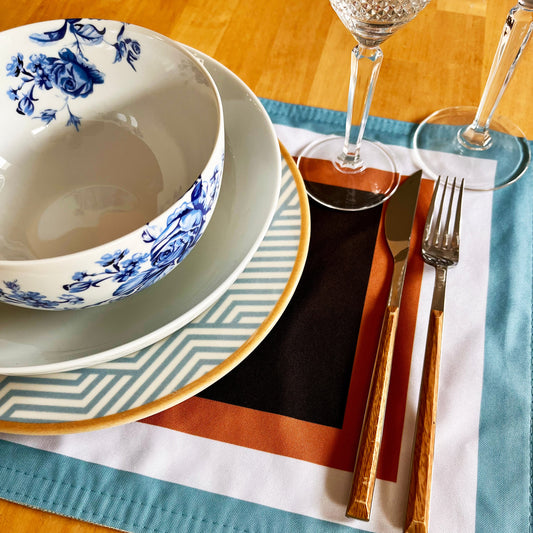 Dining table setting with blue and white floral bowl, gold cutlery, and crystal glasses on a wooden surface. Bold colours.