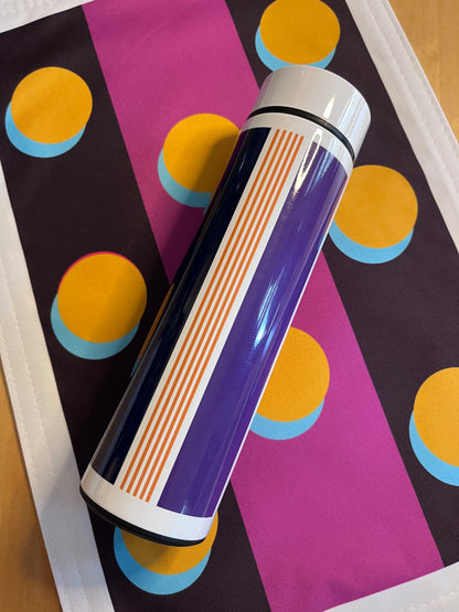 A glossy cylindrical bottle or tumbler with a purple body, orange and white stripes, and a white lid, placed diagonally on a colorful patterned placemat.