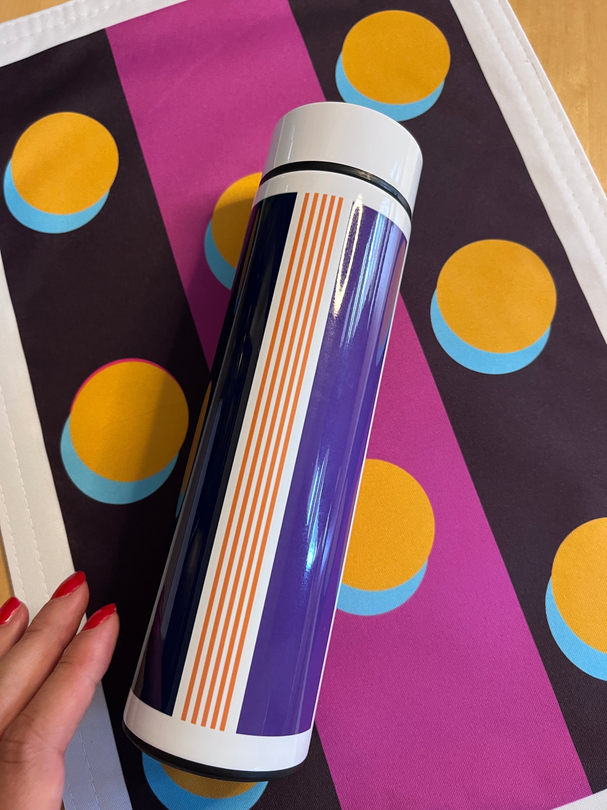 A cylindrical insulated tumbler with a purple, orange, and white striped design, held by a hand with red nail polish against a colorful patterned placemat.