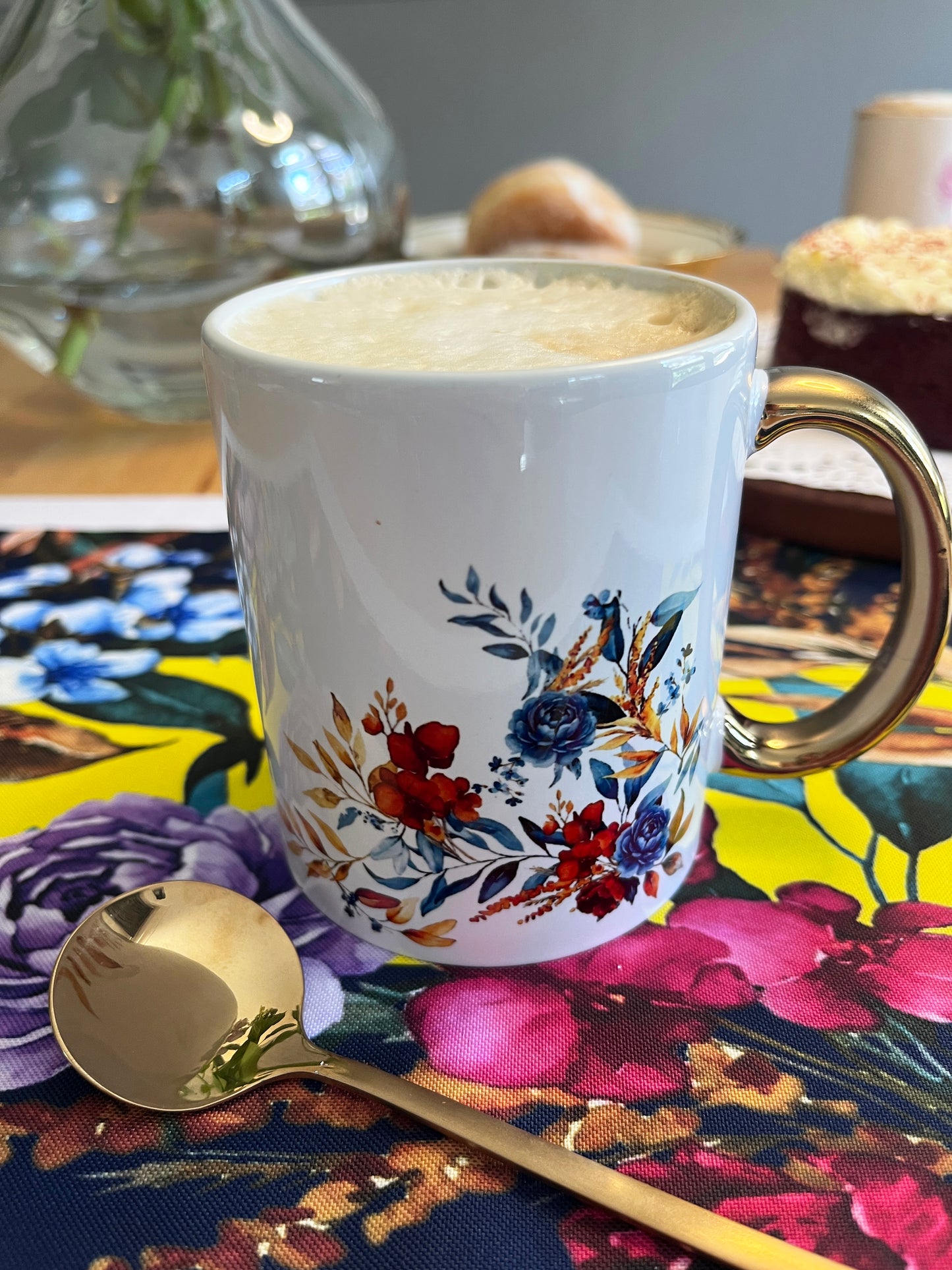A white ceramic coffee mug with a gold handle, decorated with a colorful floral pattern in red, blue, and orange. The mug is filled with a frothy hot drink and sits on a yellow and pink floral placemat next to a gold spoon.