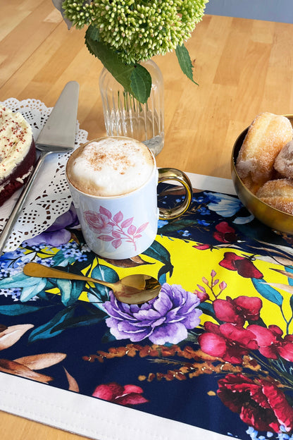A colorful floral placemat on a wooden table, styled with a white mug of cappuccino, a gold spoon, a bowl of sugar-dusted pastries, and a slice of cake.