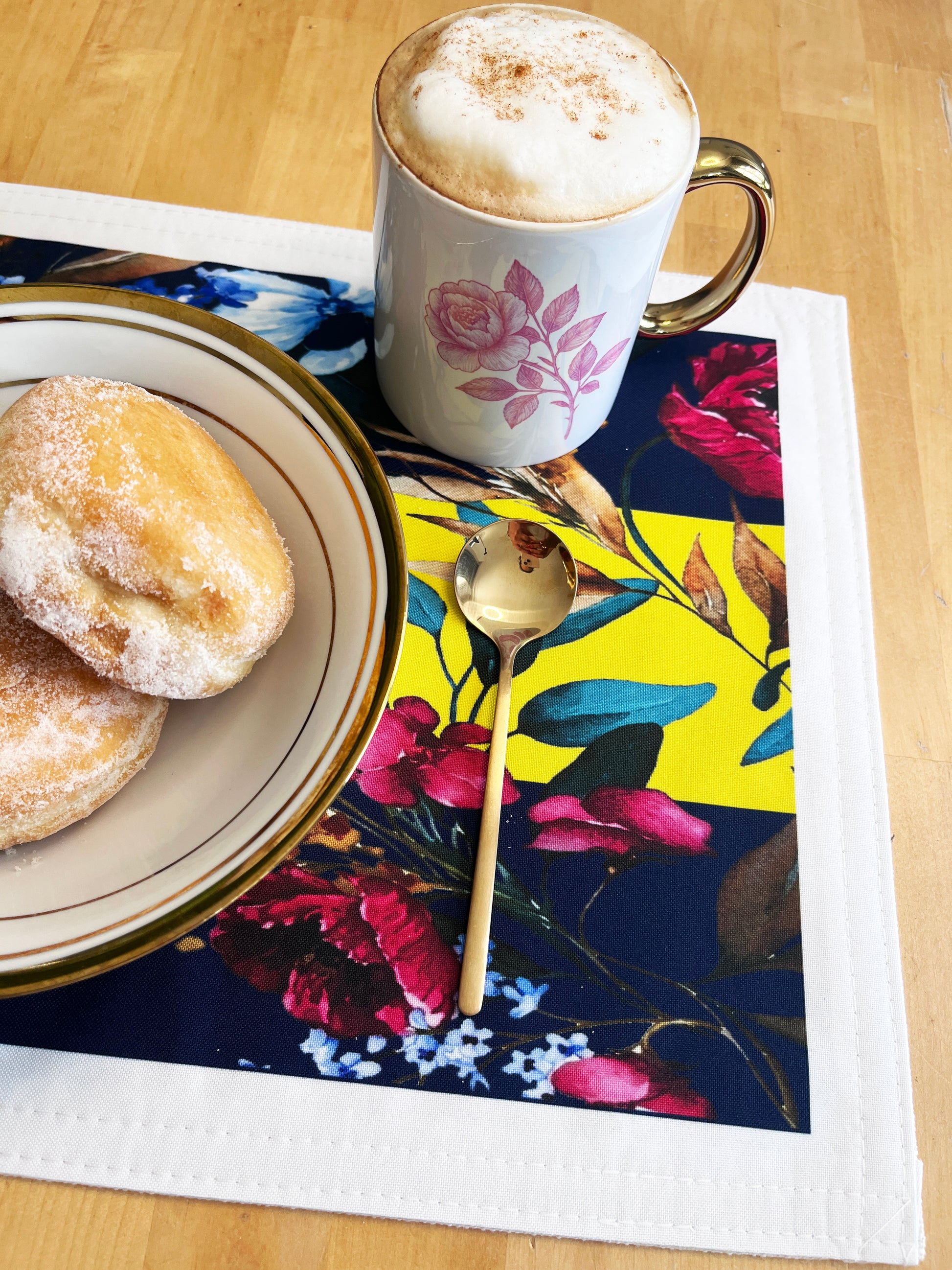 A colorful floral placemat on a light wood table styled with a white ceramic mug of cappuccino, a plate of sugar-dusted pastries, and a gold spoon.