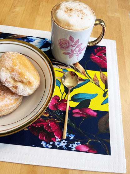 A colorful floral placemat on a light wood table styled with a white ceramic mug of cappuccino, a plate of sugar-dusted pastries, and a gold spoon.