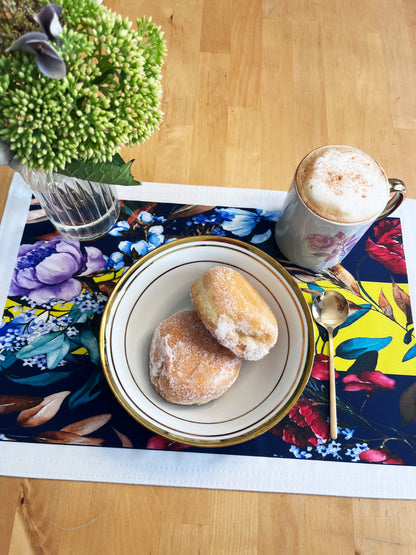 A styled breakfast table setting on a light wood table, featuring a colorful floral placemat, a white ceramic plate with two powdered pastries, a mug of cappuccino topped with cocoa powder, and a gold spoon.