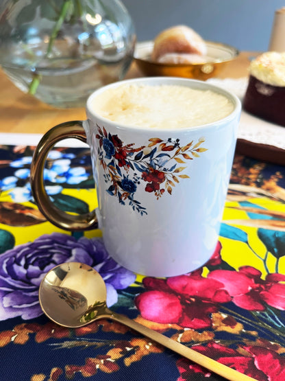 A white ceramic coffee mug with a gold handle and a colorful floral print, filled with a foamy latte, set on a vibrant floral table mat with a gold spoon in the foreground.