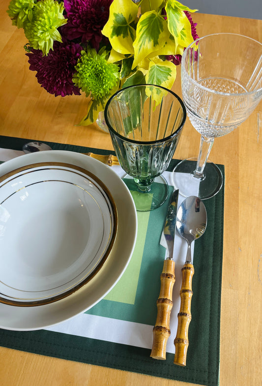 A styled tabletop place setting on a wooden table, featuring a white ceramic plate with gold trim, a green and white placemat, bamboo-handled silver cutlery, a green ribbed tumbler, a clear wine glass, and a colorful flower arrangement in the background.