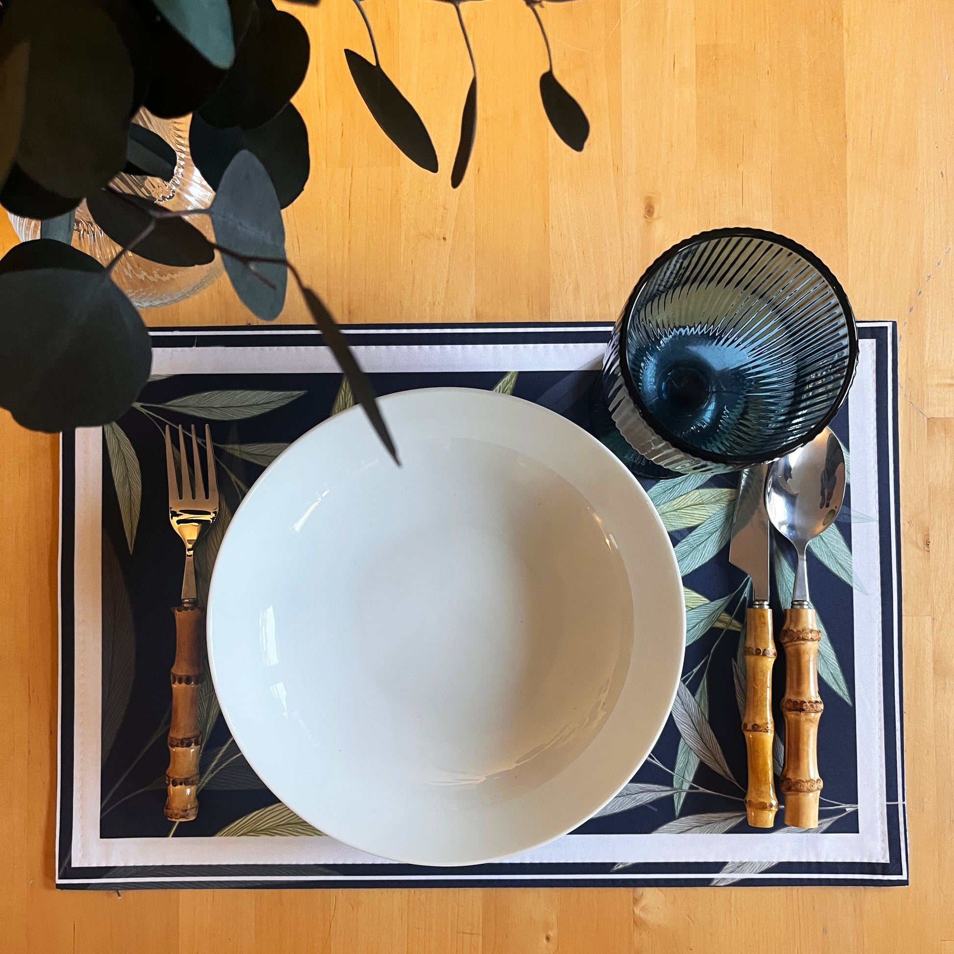 Dining setup with white bowl, black glass, bamboo cutlery on a decorative placemat, natural enviroment