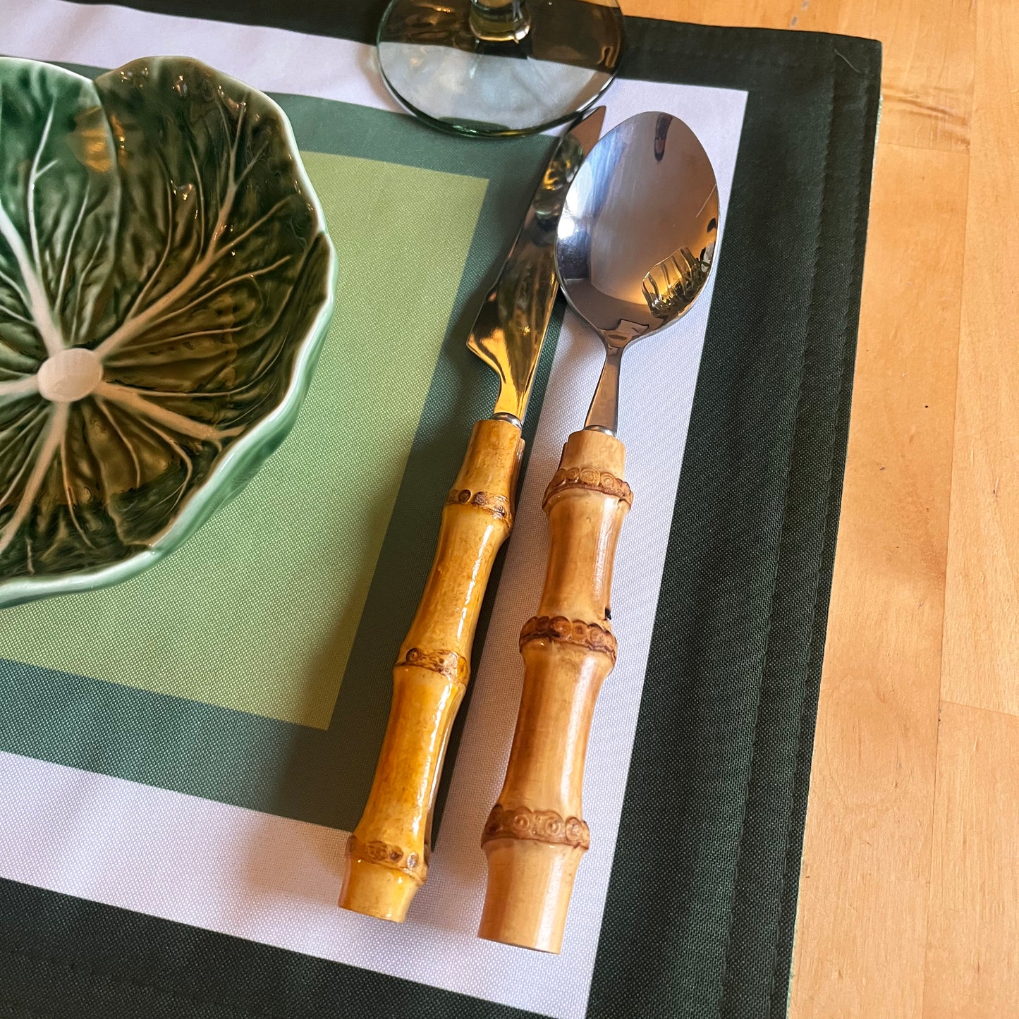 Bamboo-handled spoon and fork on a green and white placemat with a Bordallo Pinheiro ceramic leaf-patterned bowl.
