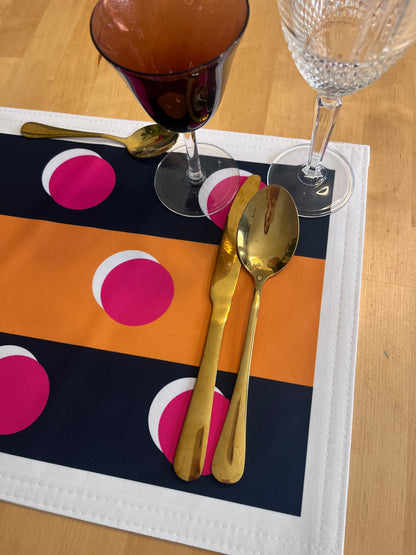 A close-up of a place setting on a wooden table, featuring a rectangular placemat with bold horizontal stripes in navy blue, orange, and white, decorated with large hot pink polka dots. Gold-toned flatware (a spoon, knife, and spoon) is arranged on the placemat, alongside two wine glasses.