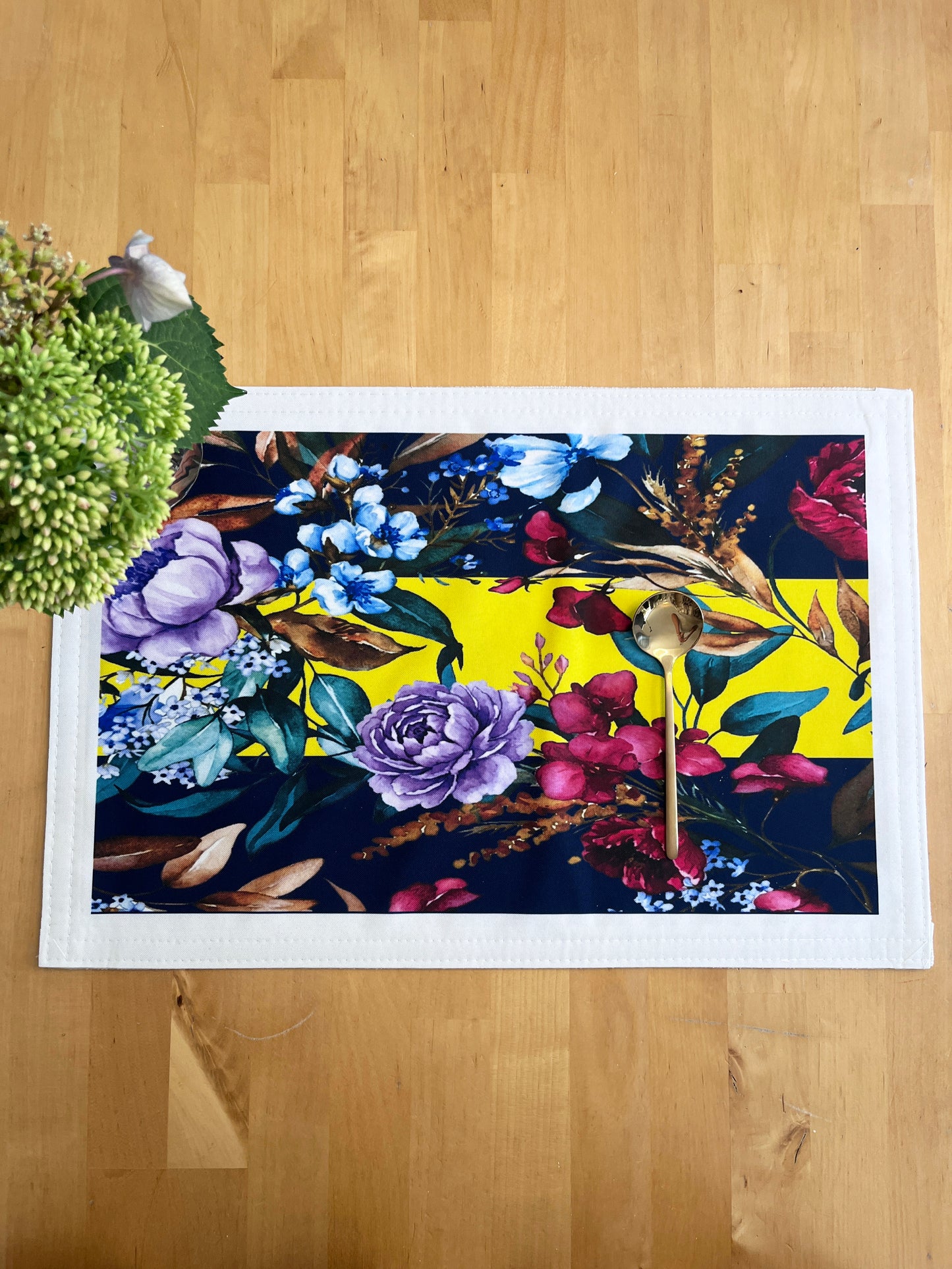 A rectangular white table runner with a vibrant floral print, laid flat on a light wood floor next to a green plant.