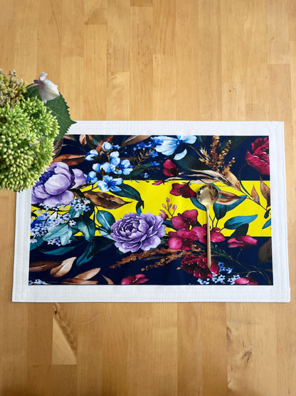 A rectangular white table runner with a vibrant floral print, laid flat on a light wood floor next to a green plant.