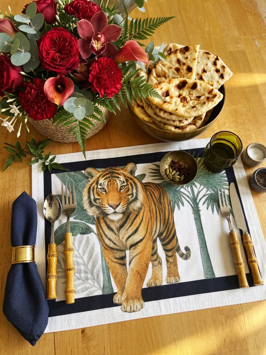A styled dining table setting featuring a rectangular placemat printed with a large tiger and palm trees, surrounded by bamboo-handled cutlery, a navy napkin, flatbread, and a floral arrangement on a wooden table.