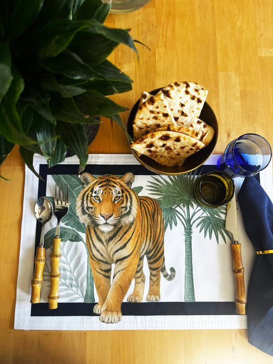 A styled tabletop setting featuring a rectangular placemat printed with a tiger and palm tree design, surrounded by cutlery with bamboo handles, folded flatbread, a blue glass, and a dark napkin on a wooden table.