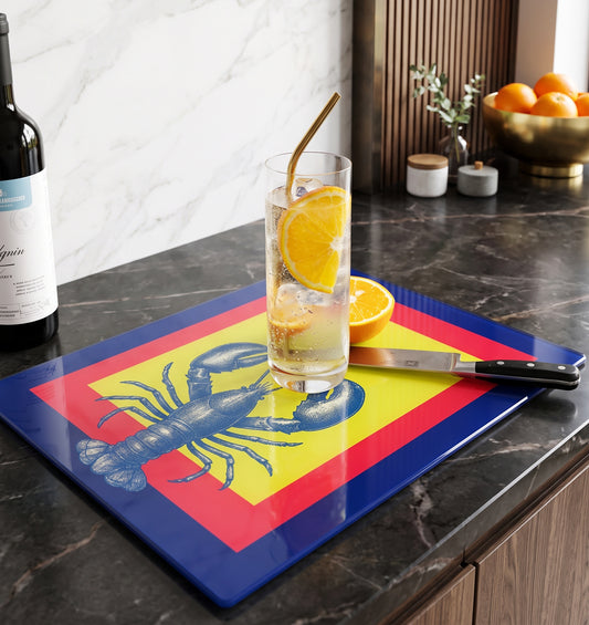 A rectangular glass cutting board with a blue lobster over a yellow and red background design, placed on a dark marble kitchen countertop. A tall glass of iced drink with orange slices, a knife, and a wine bottle are arranged on top of the board in a kitchen setting.
