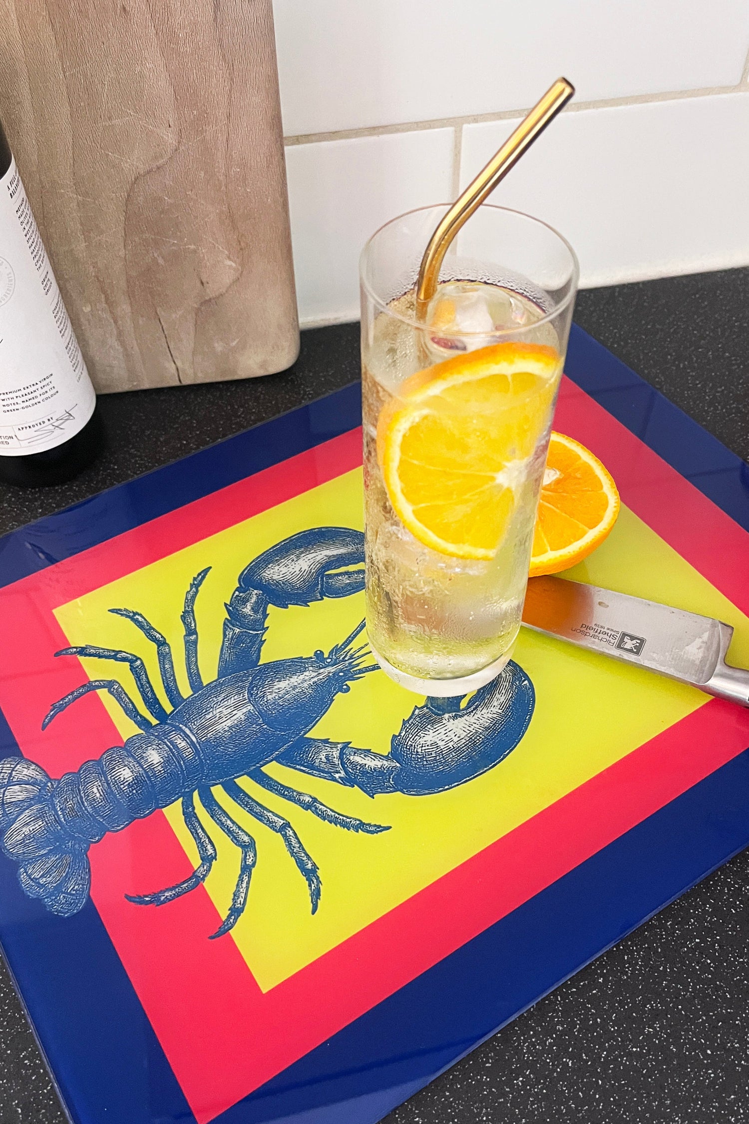 Glass of iced drink with a straw on a tray with a lobster design, next to a bottle and cutting board.