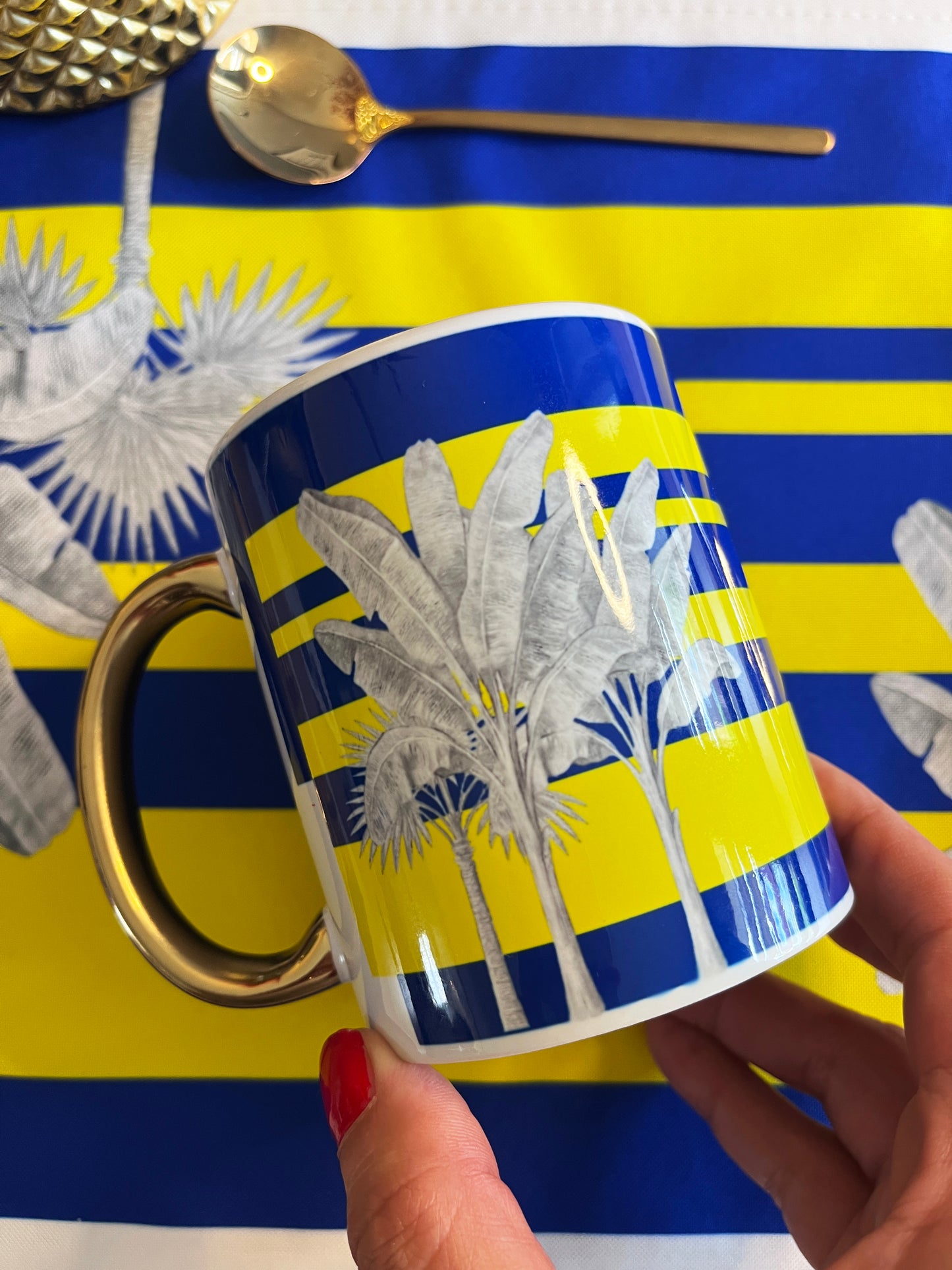 A hand with red nail polish holds a white ceramic mug featuring a blue and yellow striped tropical banana leaf pattern and a metallic gold handle. The background is a matching blue and yellow striped placemat with a gold spoon resting on it.
