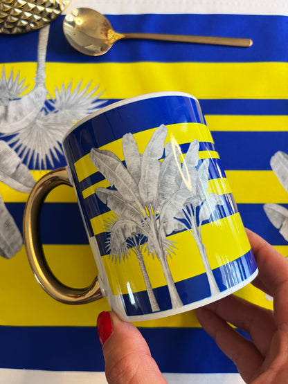 A hand with red nail polish holds a white ceramic mug featuring a blue and yellow striped tropical banana leaf pattern and a metallic gold handle. The background is a matching blue and yellow striped placemat with a gold spoon resting on it.