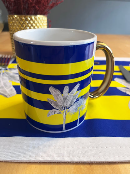 A ceramic mug with a blue and yellow striped pattern, featuring a white palm tree illustration and a metallic gold handle, set on a matching blue and yellow placemat.