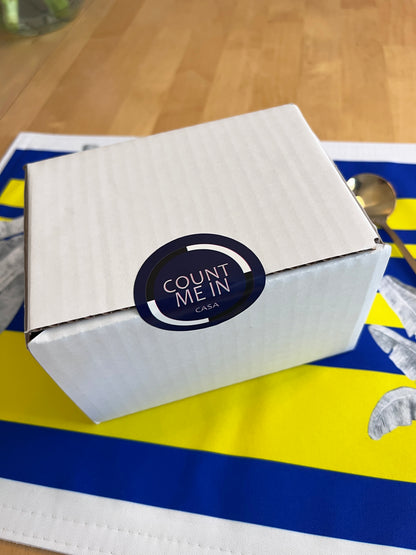 A white cardboard box with a circular black and white 'COUNT ME IN CASA' sticker on top, resting on a blue and yellow striped surface with a spoon nearby, on a wooden table.