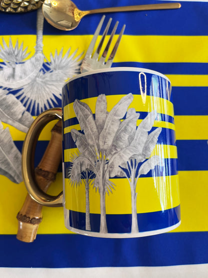 A ceramic mug with a blue and yellow striped design and white palm tree illustrations, styled with gold-toned cutlery on a matching blue and yellow striped background.