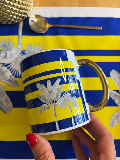 A hand with red nail polish holds a white ceramic mug featuring a blue and yellow striped pattern with silver palm tree illustrations. The background is a matching blue and yellow striped placemat with a gold spoon and textured gold object partially visible.
