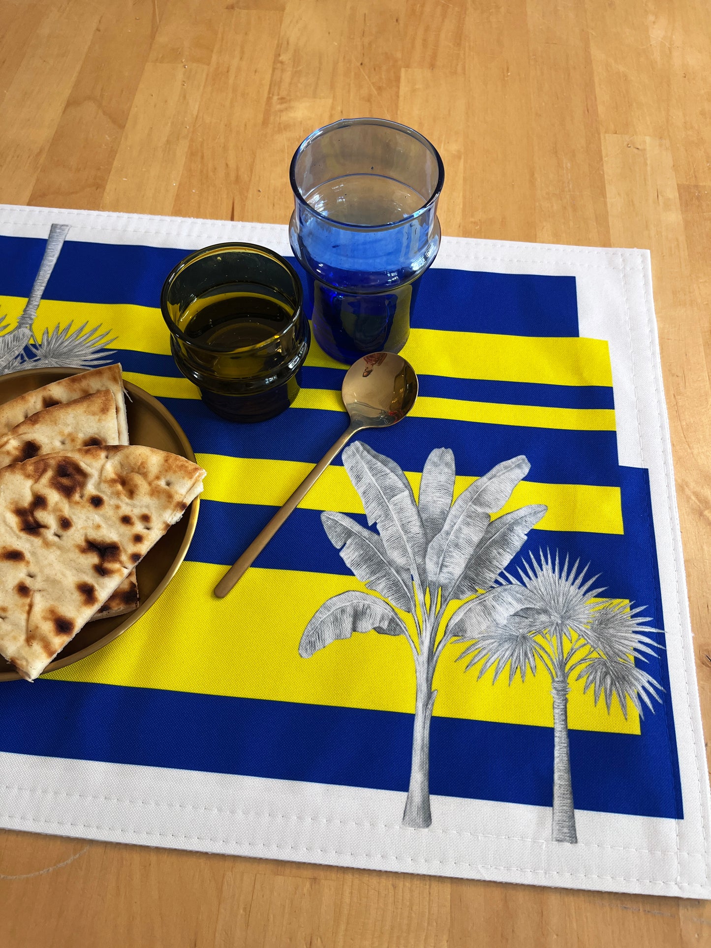 A rectangular woven placemat with bold blue and yellow horizontal stripes, white border, and silver palm tree illustrations, styled on a light wood table with folded flatbread, stacked drinking glasses, and a spoon.
