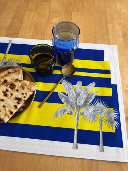 A rectangular woven placemat with bold blue and yellow horizontal stripes, white border, and silver palm tree illustrations, styled on a light wood table with folded flatbread, stacked drinking glasses, and a spoon.