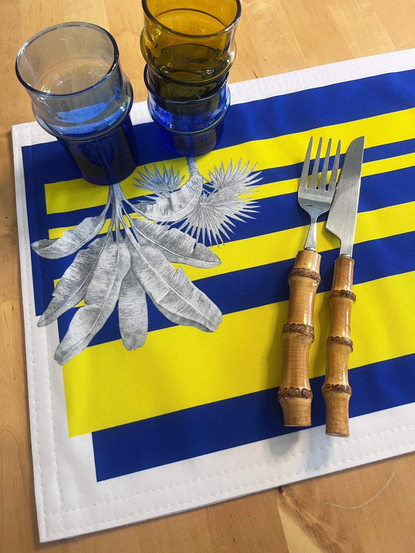 A square table mat featuring the flag of Suriname, styled on a wooden table with stacked blue and amber glass tumblers, a silver leaf print, and a bamboo-handled fork and knife.