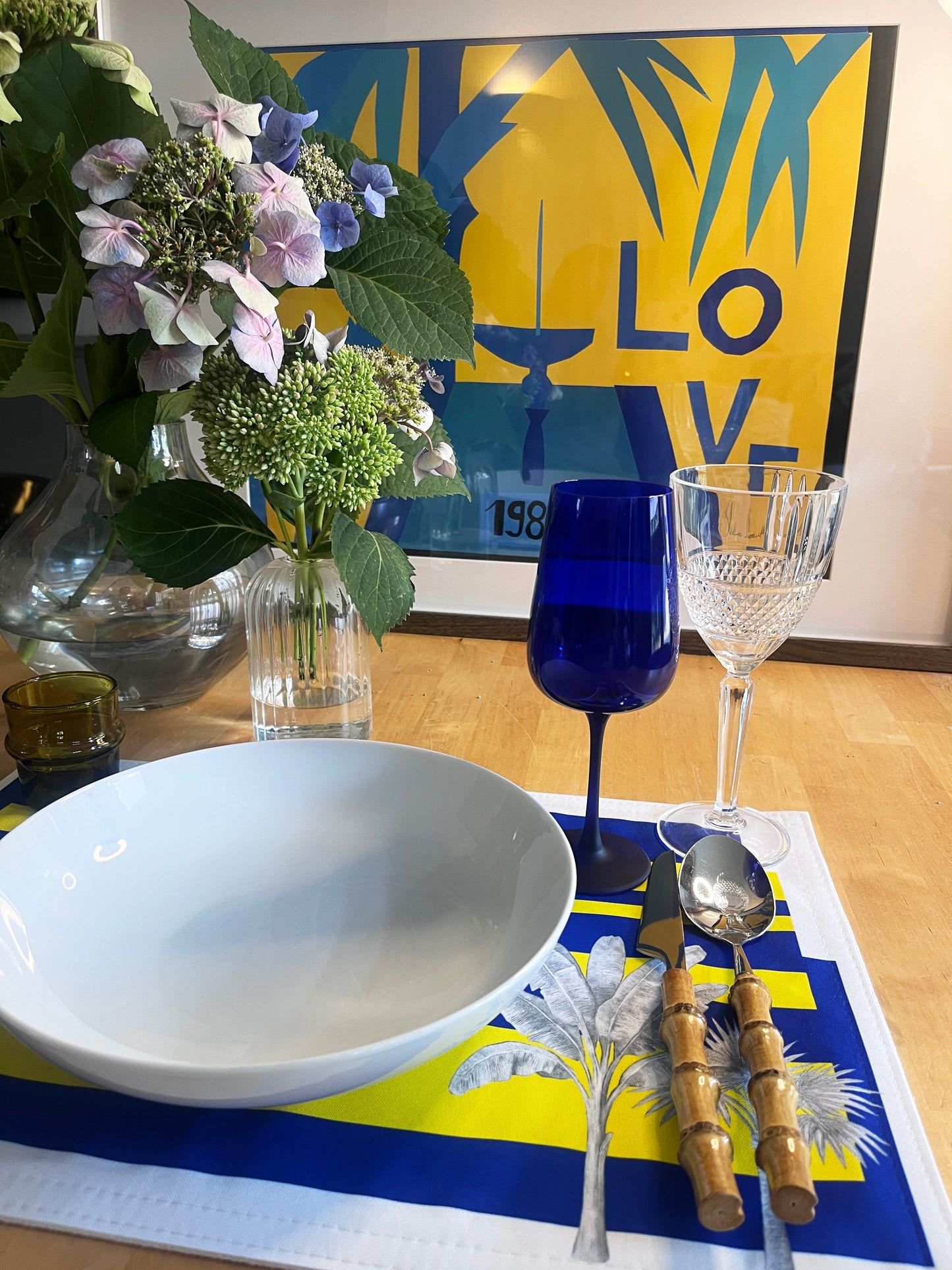 A styled dining table setting featuring a large white ceramic bowl, a cobalt blue wine glass, a clear textured wine glass, and bamboo-handled silver cutlery on a blue and yellow placemat. A glass vase of hydrangeas and a colorful framed art print are visible in the background on a light wood table.