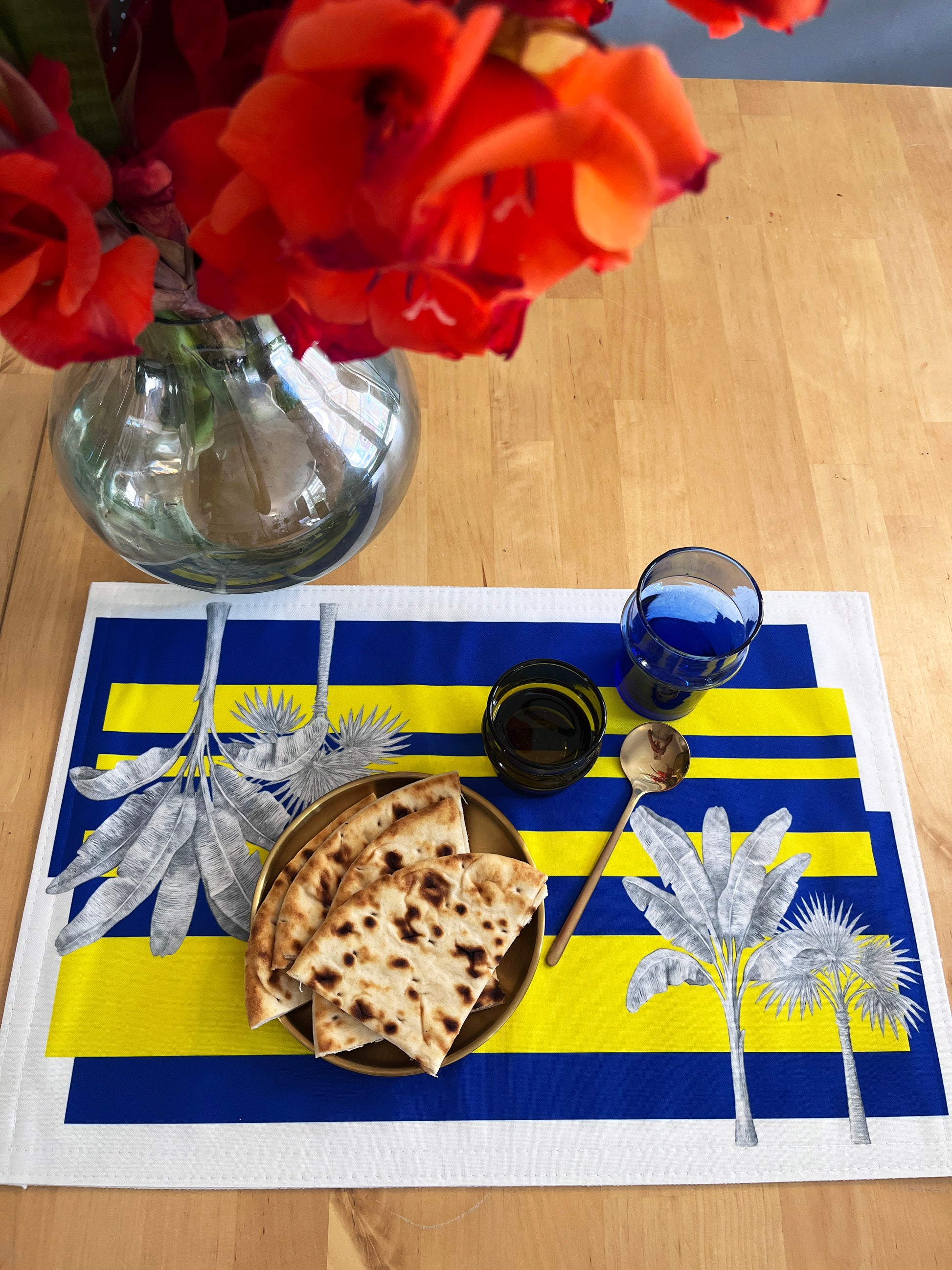 A rectangular table placemat with bold blue and yellow horizontal stripes, white borders, and silver palm tree illustrations, styled on a wooden table with a plate of flatbread, a blue glass, a spoon, and a vase of red flowers.