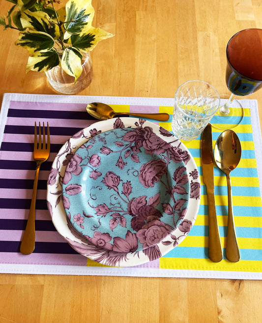 Dinner setting with floral plate, cutlery, and striped  purple, light blue and yellow, placemat on a wooden table.