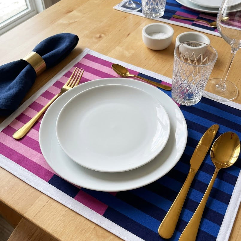Dining table setting with white plates, gold cutlery, and striped placemats.